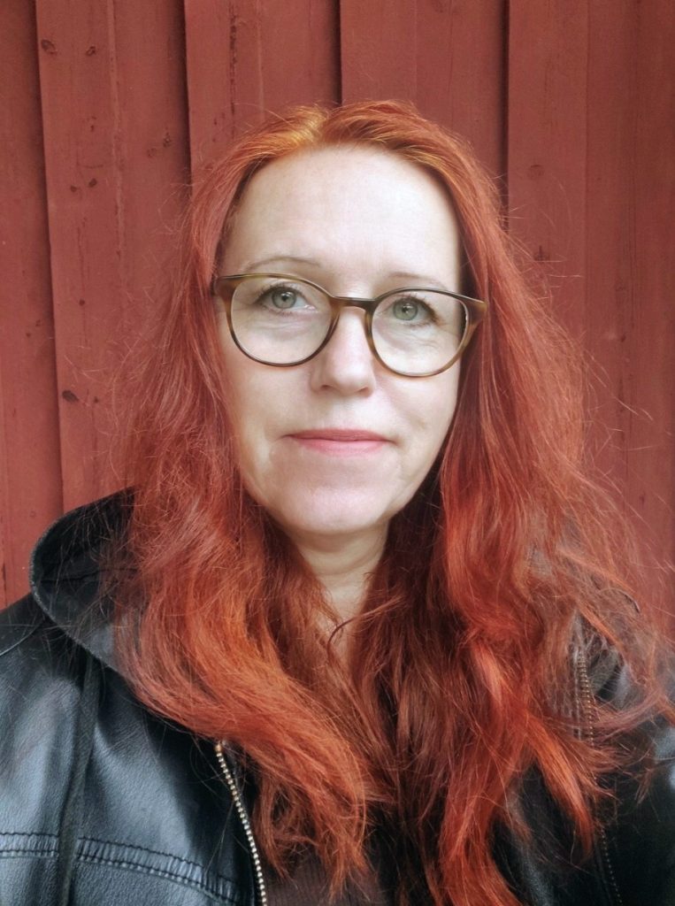 Kvinna med långt rött hår och glasögon, klädd i svart jacka, står framför en röd trävägg.