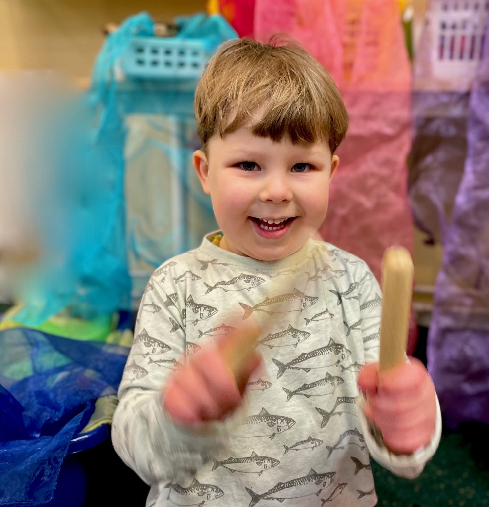 Leende barn i skjorta med fiskavtryck leker med träpinnar i ett färgglatt klassrum.