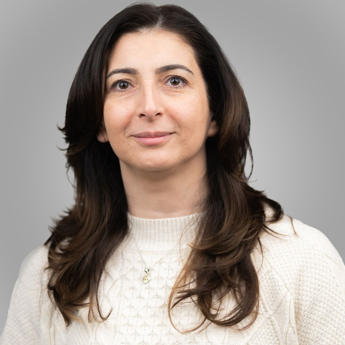 Kvinna med långt brunt hår, vit tröja och ett mjukt leende mot en grå bakgrund.