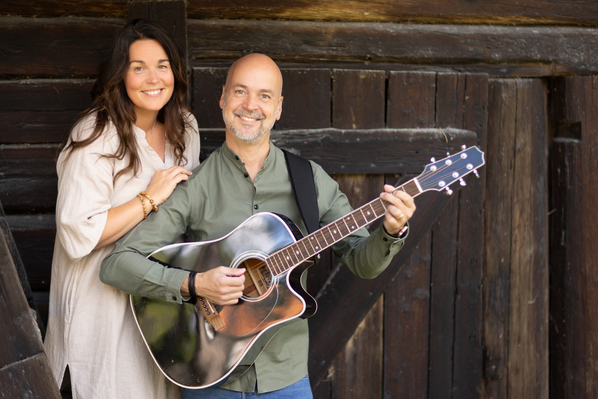 Leende man spelar gitarr medan kvinna står bredvid honom, båda framför en rustik trävägg.