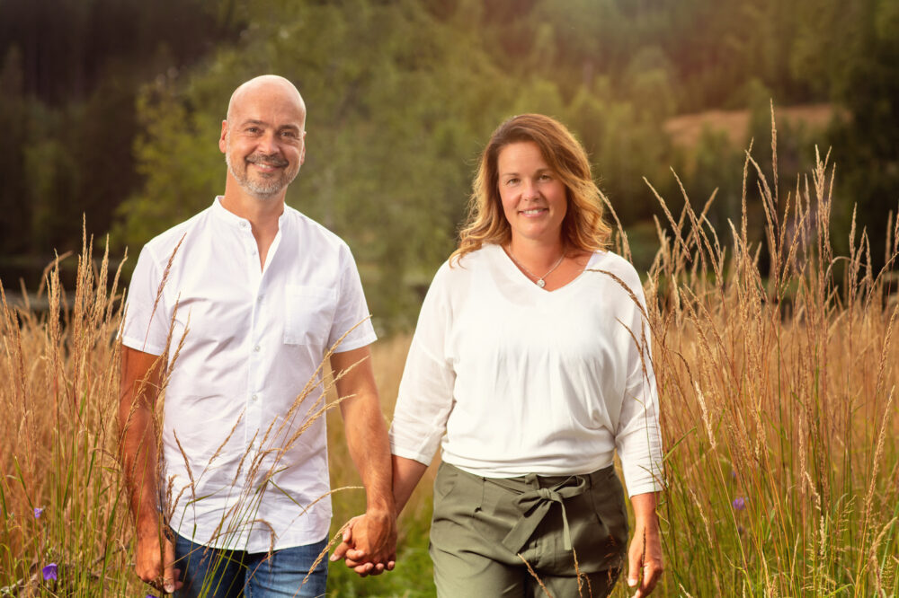 Ett leende par som håller varandra i handen står på ett fält med högt gräs och träd i bakgrunden.