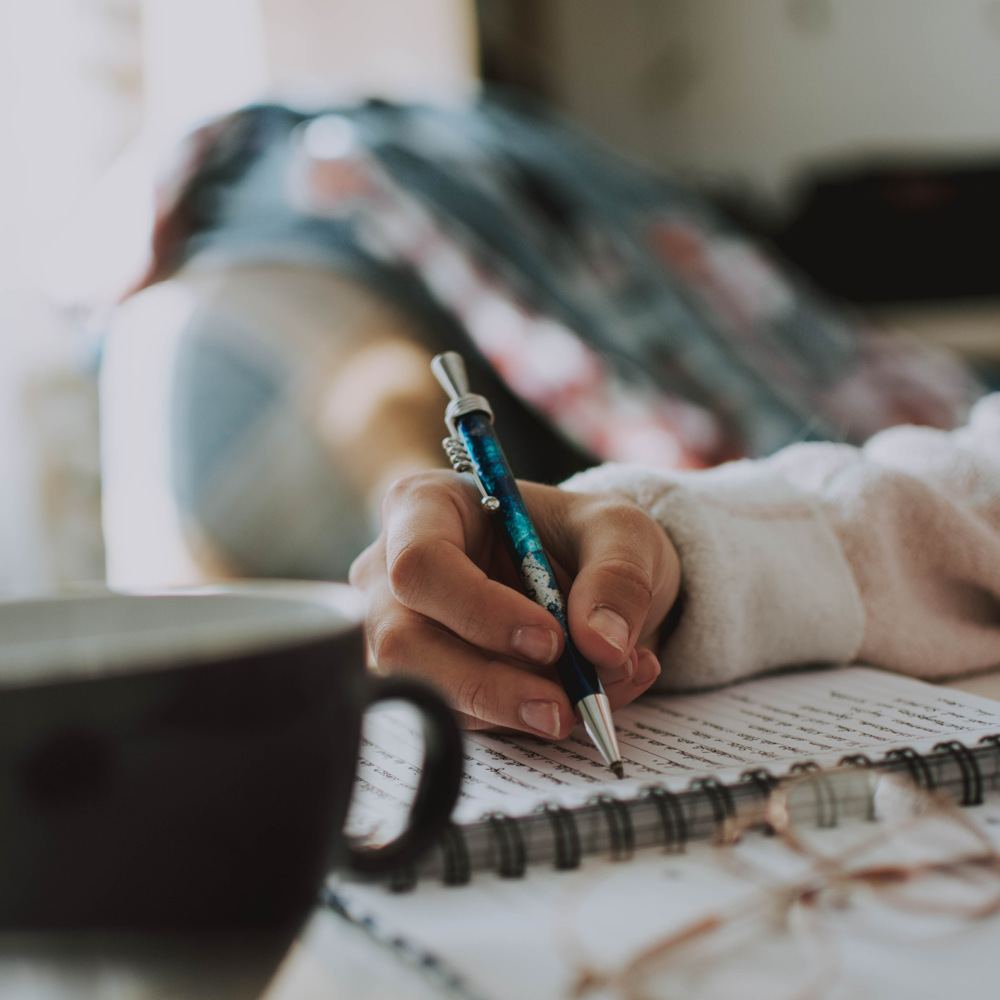 En person skriver i en anteckningsbok med en blå penna, en kaffekopp och glasögon står på bordet.