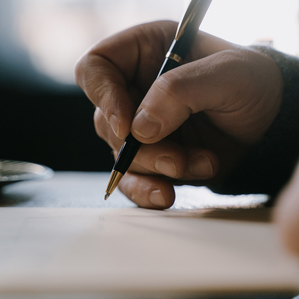 En person som håller i en penna och skriver på ett papper vid ett skrivbord.