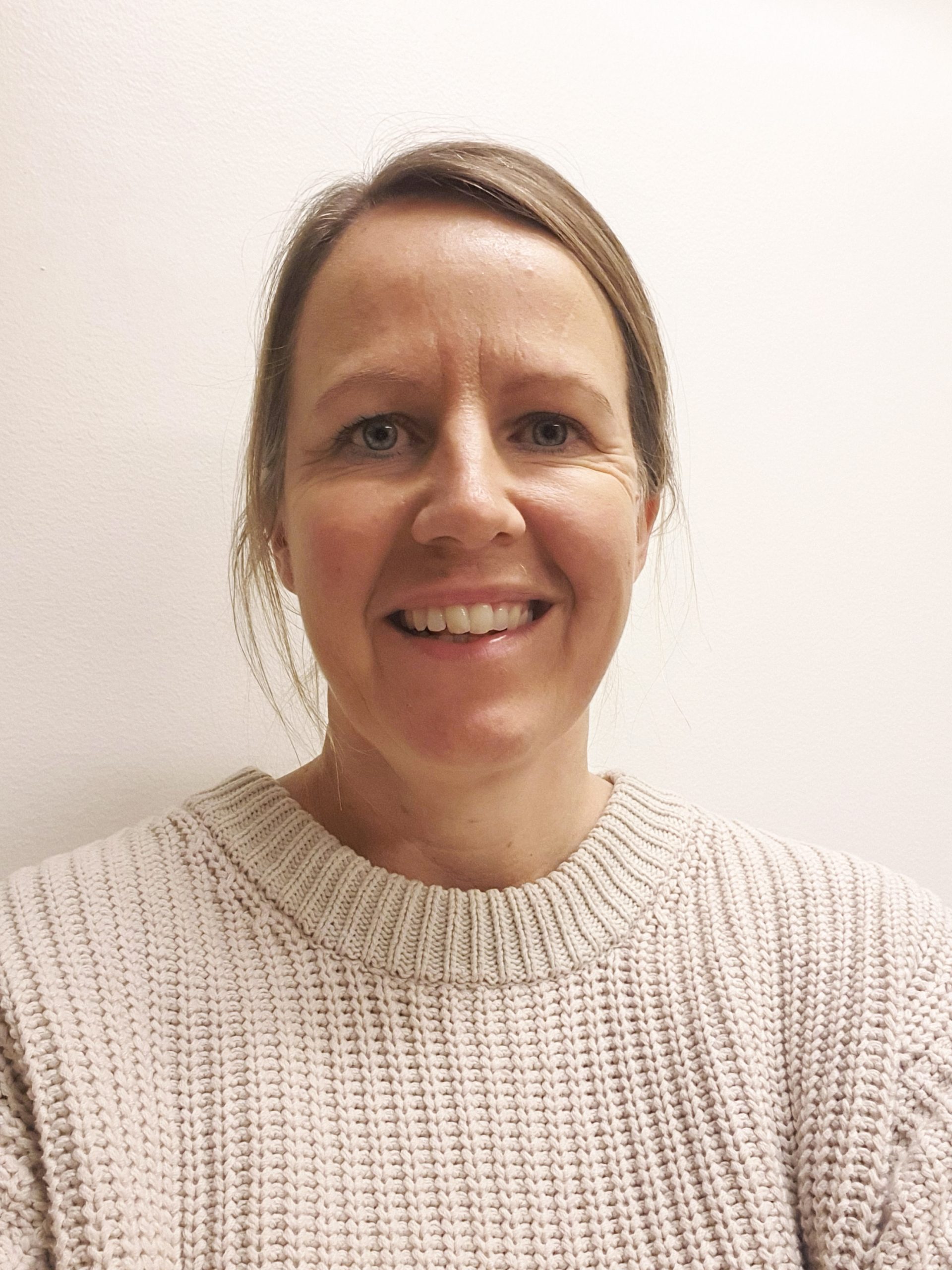 En kvinna med blont hår i en knut, klädd i en gräddvit stickad tröja, ler mot kameran mot en vit bakgrund.