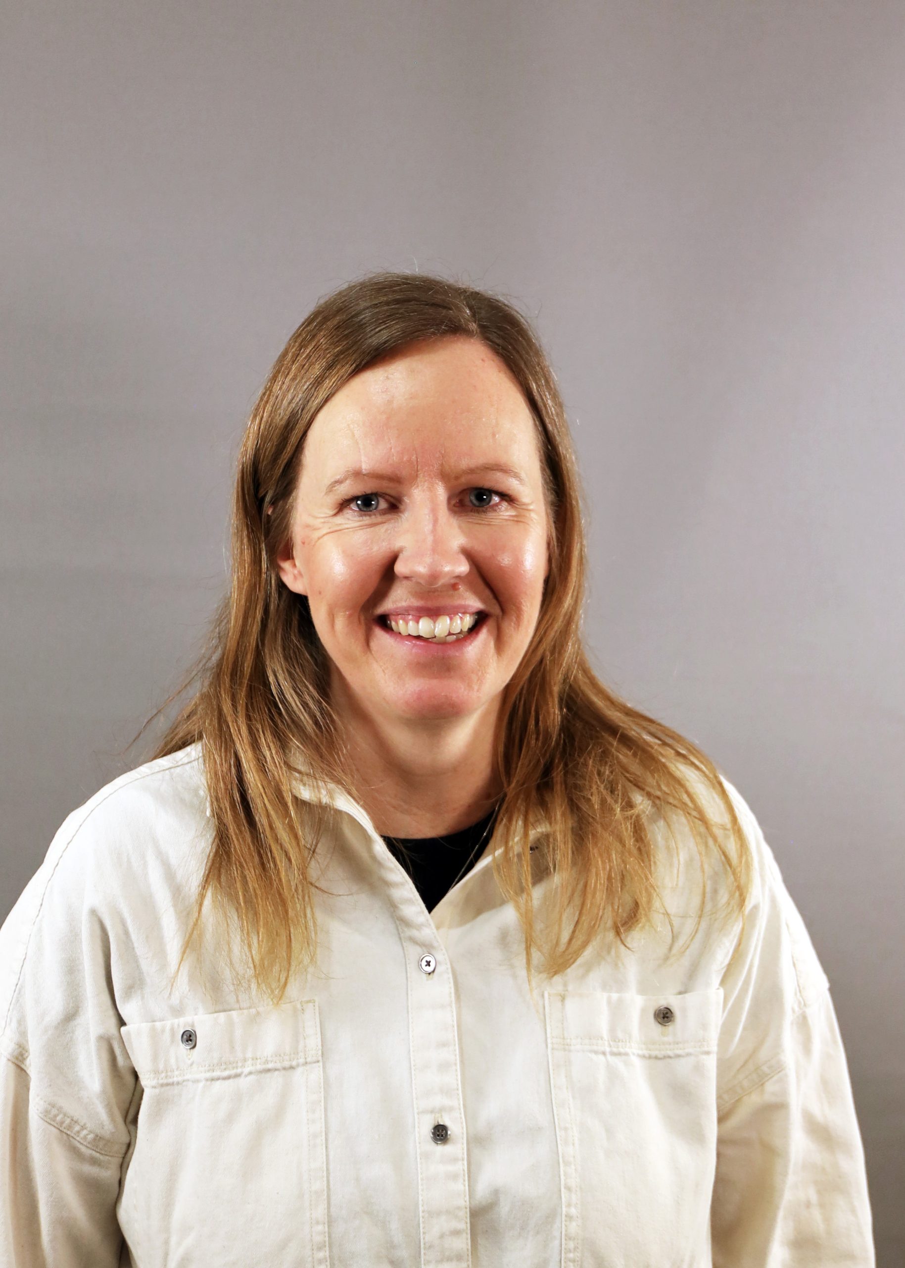 En kvinna med långt brunt hår ler, klädd i vit skjorta med knappar mot en enfärgad grå bakgrund.