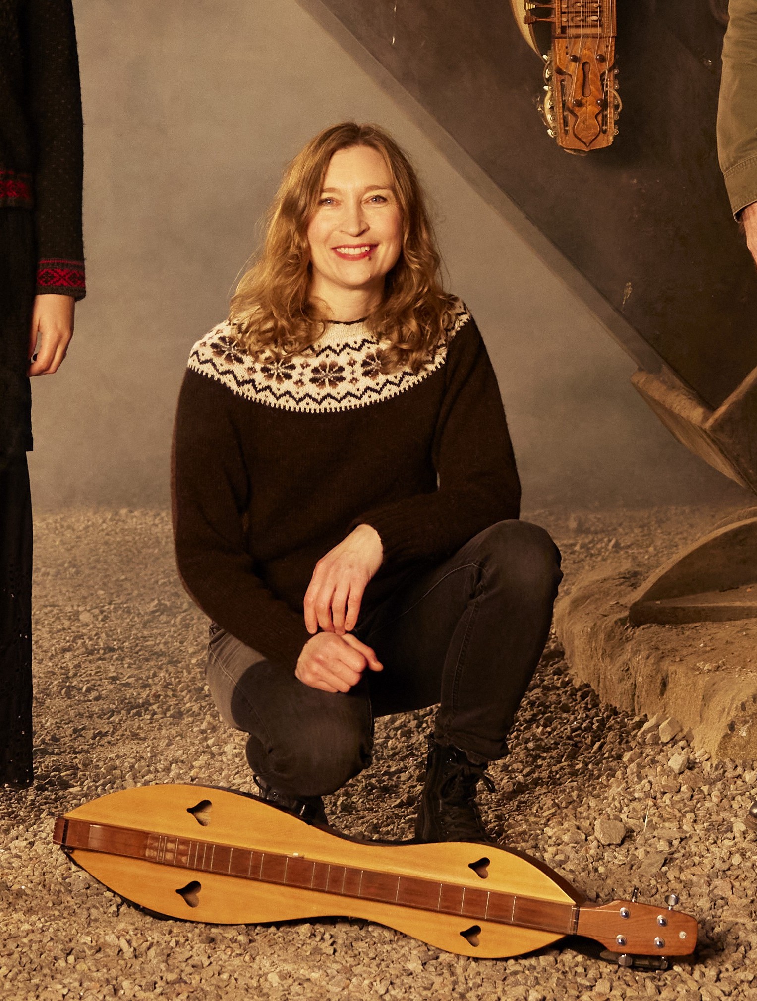 En kvinna i mönstrad tröja står på knä i gruset bredvid ett stränginstrument i trä och ler.