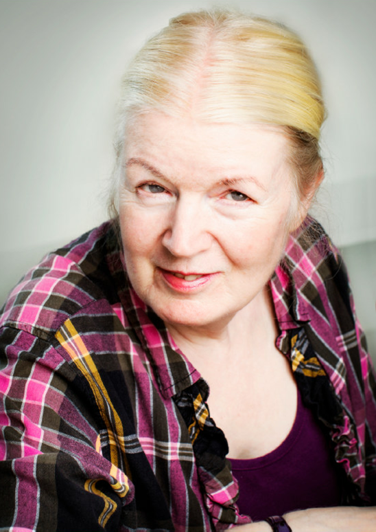 Äldre kvinna med ljust hår som är bakåtkammat, klädd i rutig skjorta och ler mot kameran.