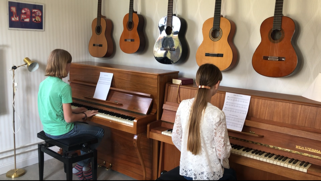 Två personer spelar piano i ett rum med sex gitarrer hängande på väggen ovanför dem.