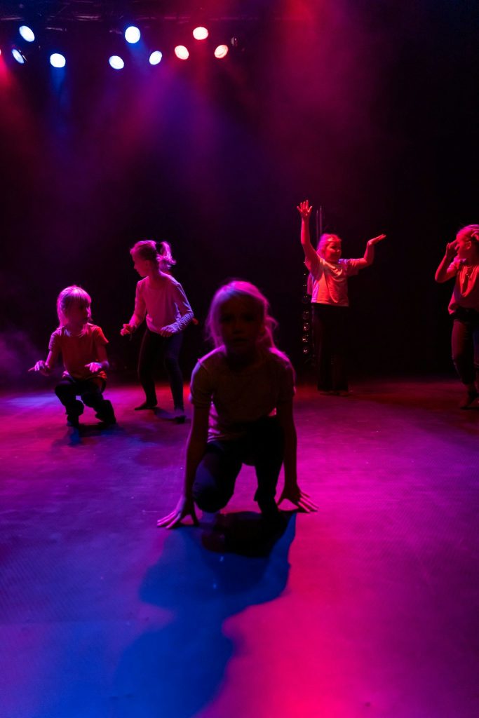 Fem barn dansar och poserar på en scen under dramatisk röd och lila belysning.