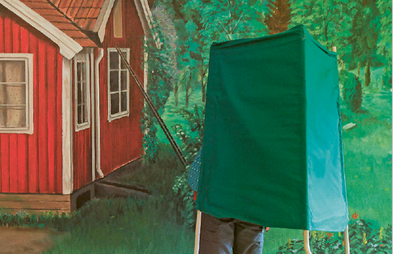 En person står i en grön röstningsbås bredvid ett rött trähus med en grönskande bakgrund.