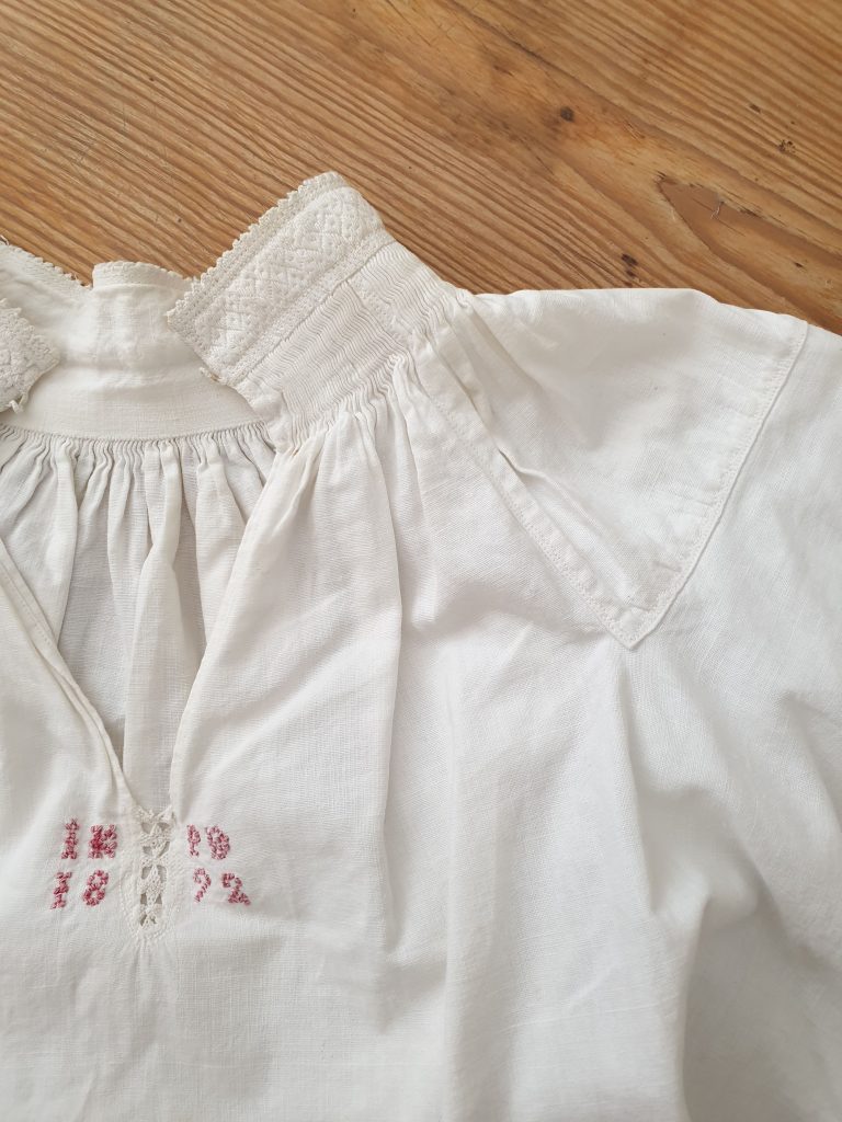 Vit folkdräktskjorta med spetskrage och rosa broderi, placerad på en träyta.