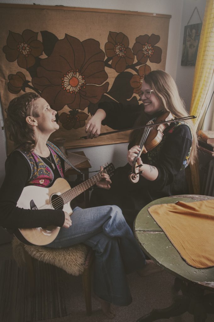 Två kvinnor ler och spelar gitarr och fiol tillsammans i ett mysigt rum i vintagestil med blommig inredning.