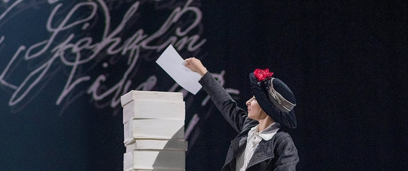 En person i tidstypisk dräkt håller upp ett papper bredvid en hög med vita lådor mot en mörk bakgrund.