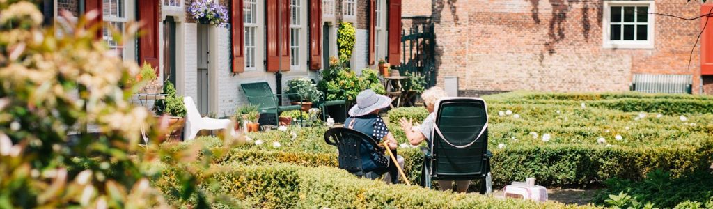 Två personer sitter i trädgårdsstolar och pratar på en prydligt klippt innergård framför tegelhus.