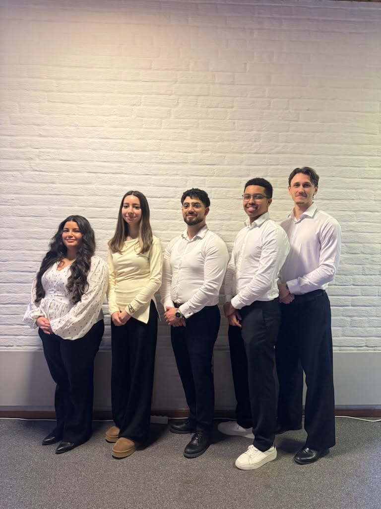 Fem personer klädda i vardagskläder står sida vid sida mot en vit tegelvägg och ler.