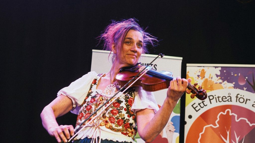 En kvinna i traditionell klädsel spelar fiol på scenen med färgglada banderoller i bakgrunden.