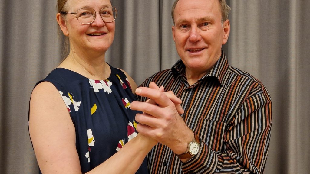 Två leende vuxna håller varandra i handen och poserar tillsammans framför en grå gardin.