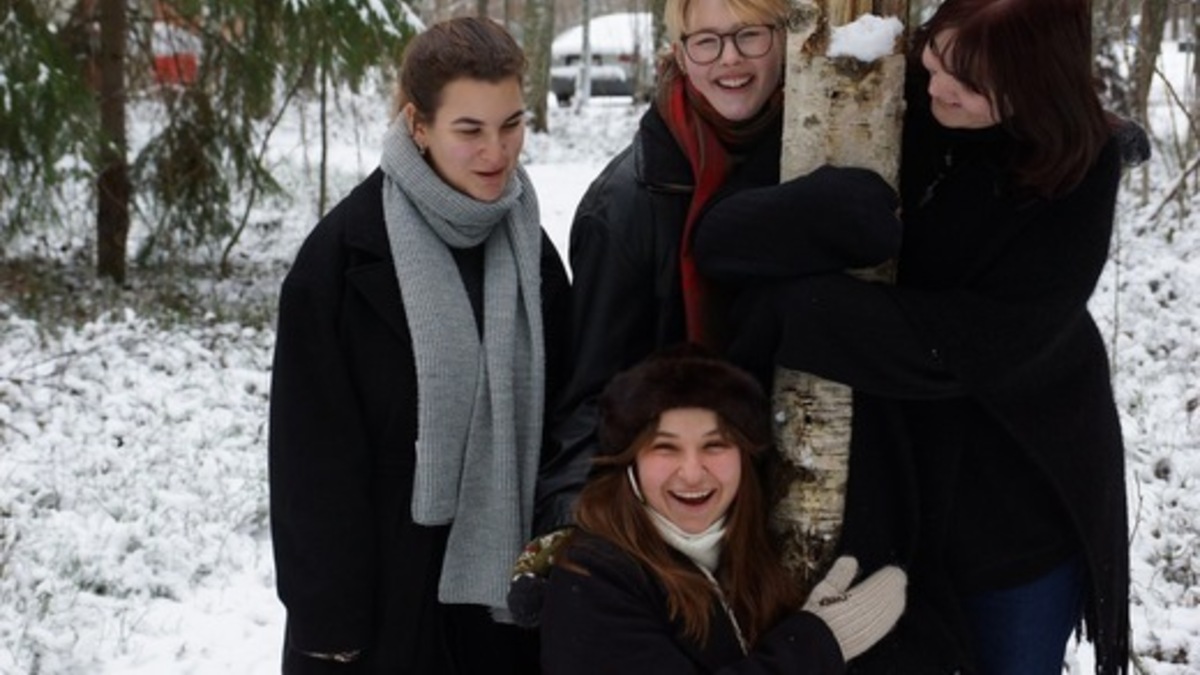 Fyra personer i vinterjackor och halsdukar ler och kramar ett träd i en snöig skog.