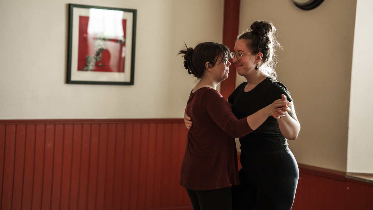 Två kvinnor håller varandra i handen och dansar tillsammans i ett varmt upplyst rum med röda paneler och inramad konst på väggen.