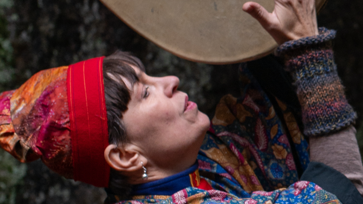 Person med färgglad klädsel och röd huvudduk, håller en trumma över huvudet, utomhus.