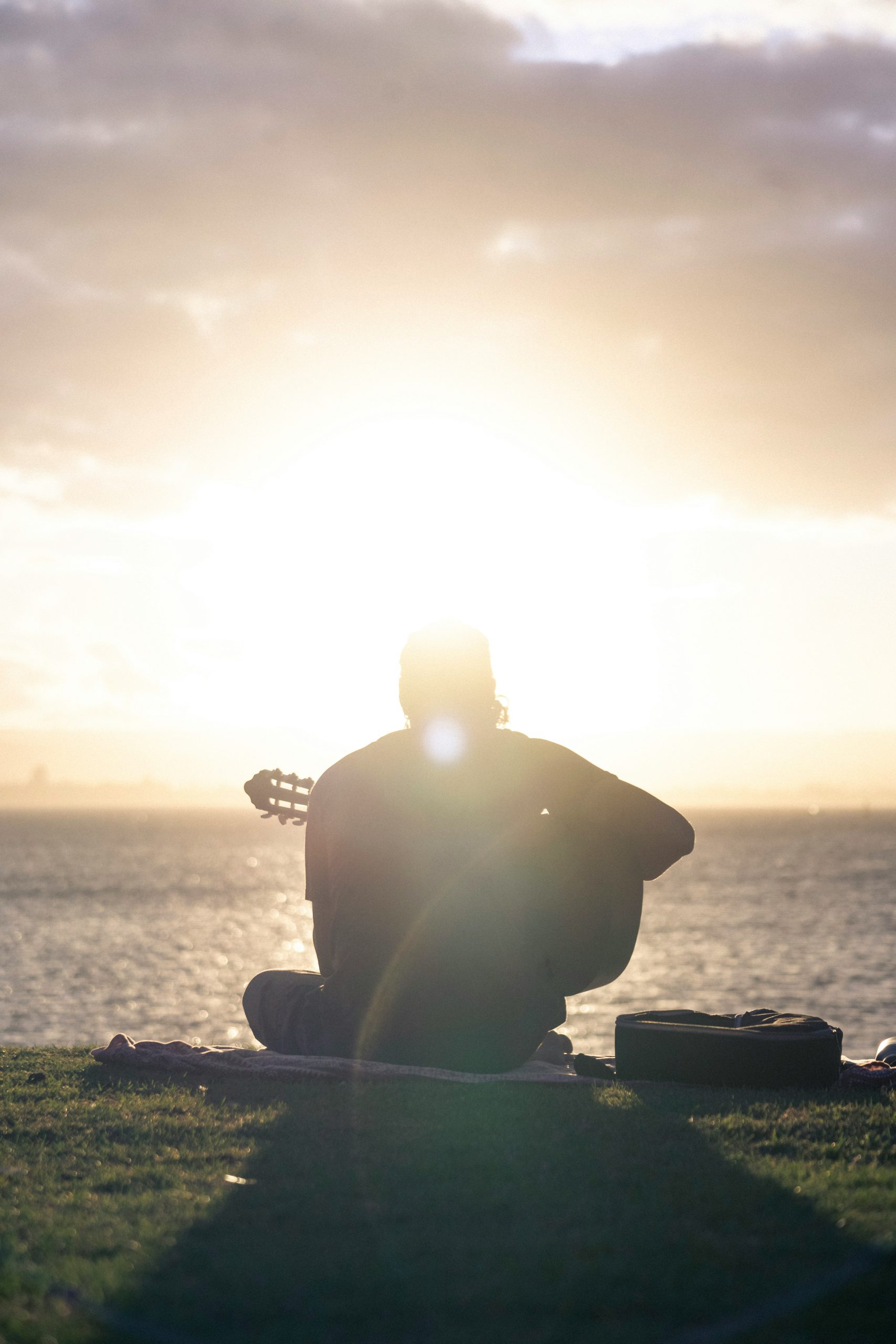 Silhuett av en person som spelar gitarr vid vattnet i solnedgången, med ett gitarrfodral på gräset i närheten.