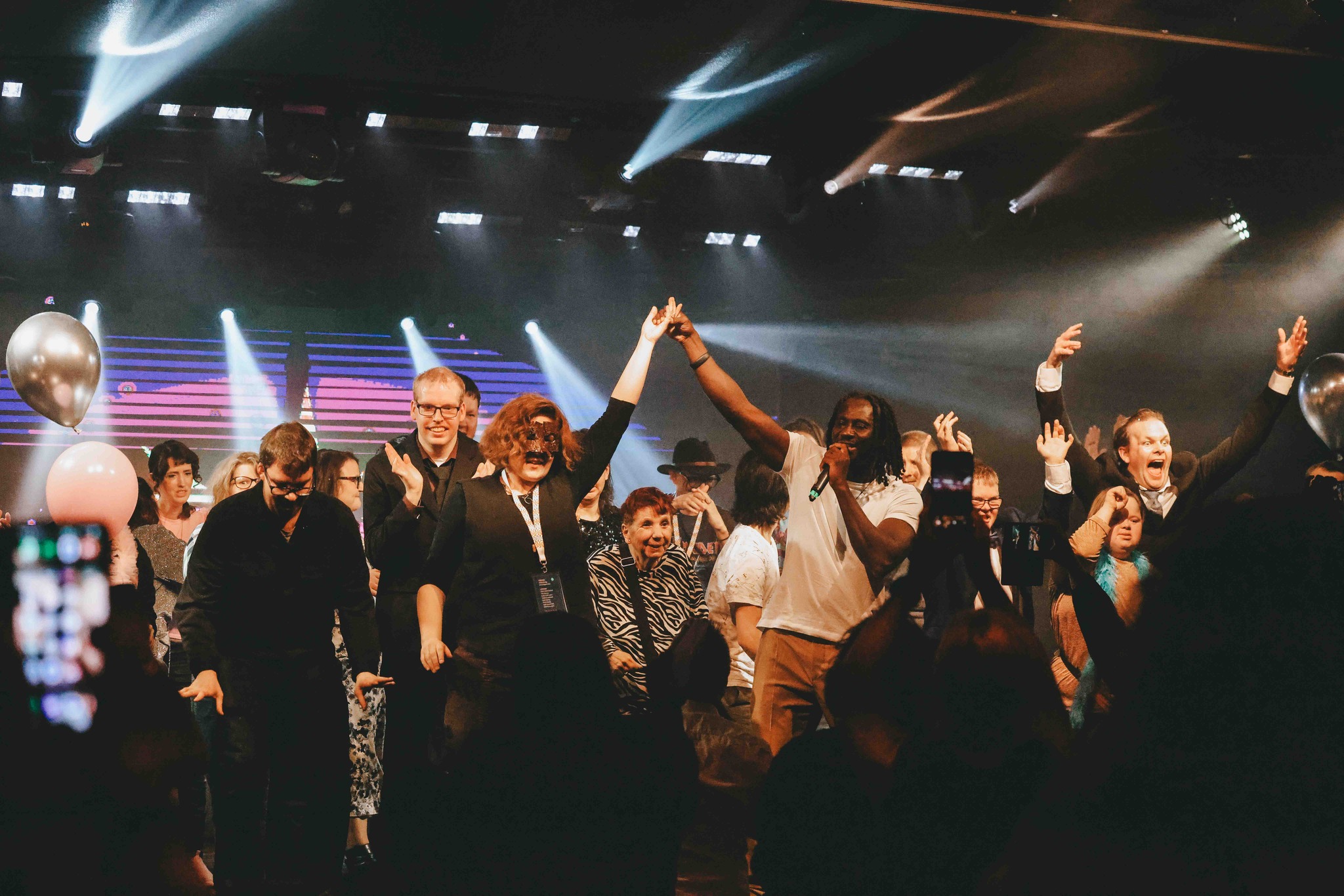 En grupp människor på scenen firar tillsammans, ler och lyfter sina händer under färgglada lampor.