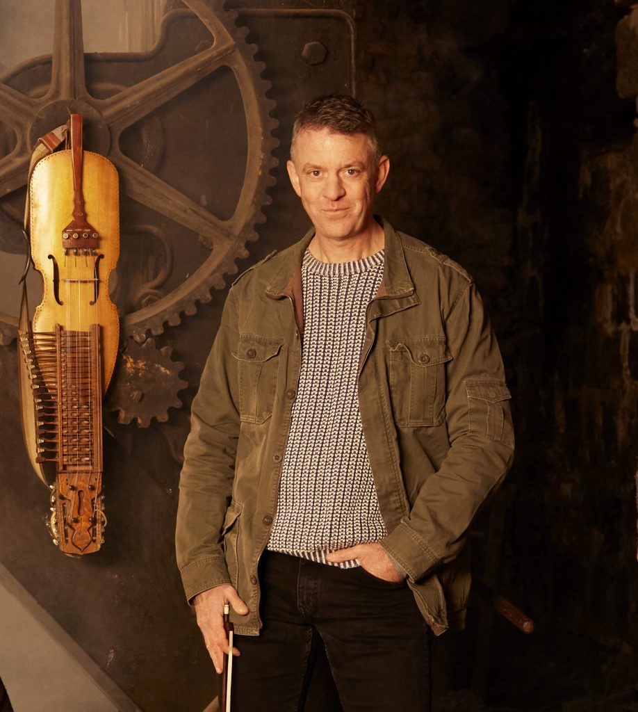En man i ledig kavaj står bredvid en nyckelharpa som är monterad på en vägg med stora mekaniska kugghjul.