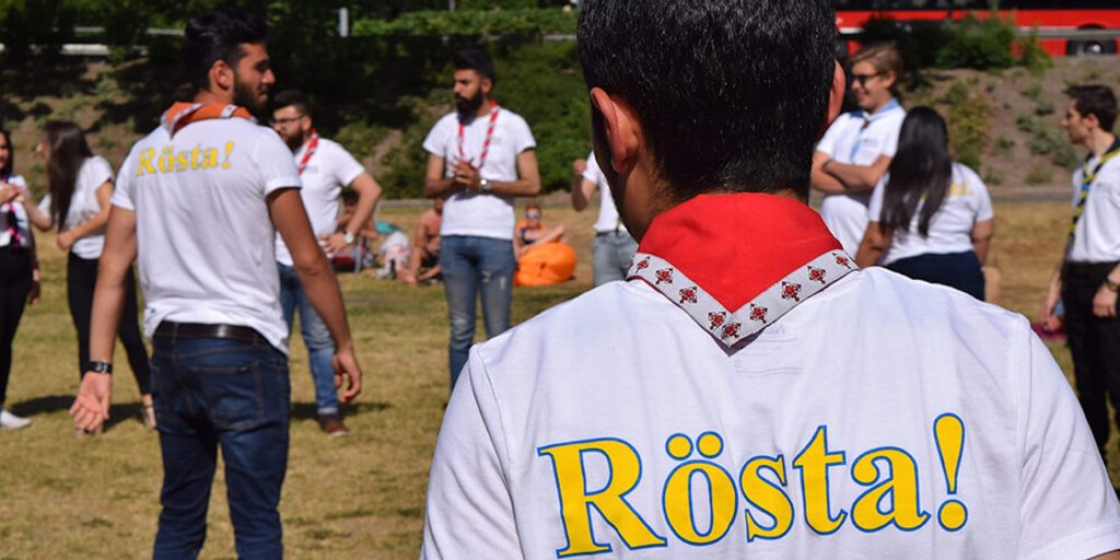 Personer i vita tröjor med texten "Rösta!" samlas utomhus på en gräsmatta i en grupp, några vända mot varandra.