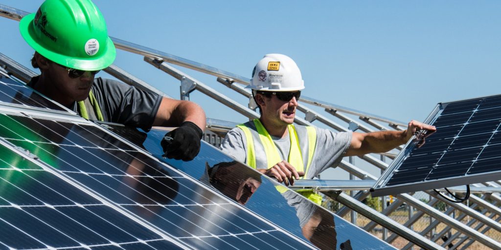Två arbetare i skyddshjälm installerar solpaneler utomhus en solig dag.