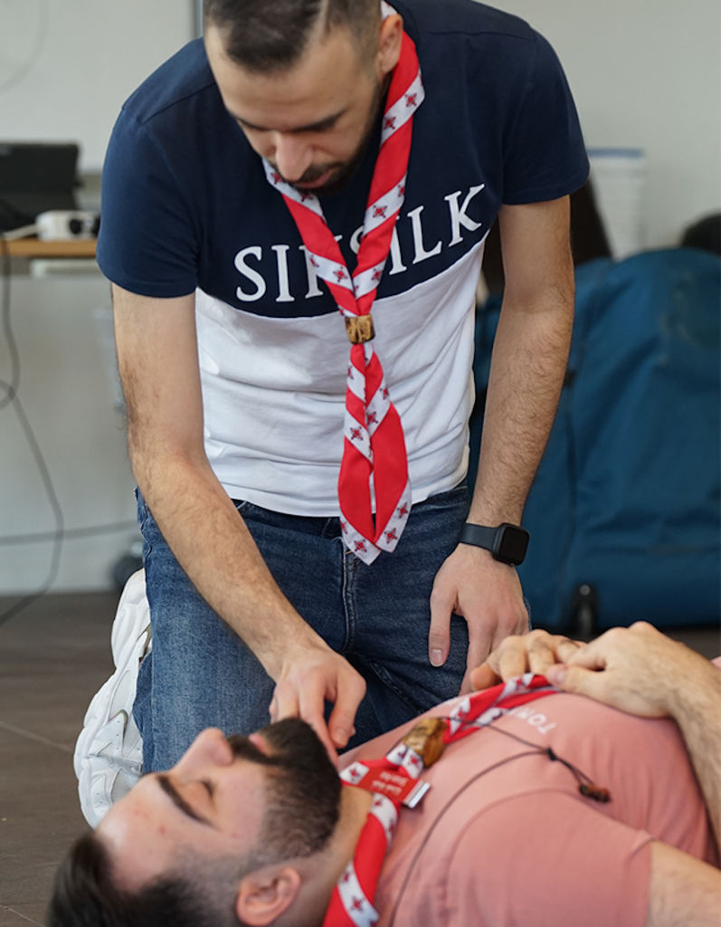 En man kontrollerar en annan mans andning under en utbildning i första hjälpen; båda bär röda scouthalsdukar.