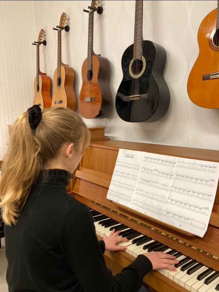 En flicka spelar piano framför noter, med flera gitarrer hängande på väggen bakom henne.