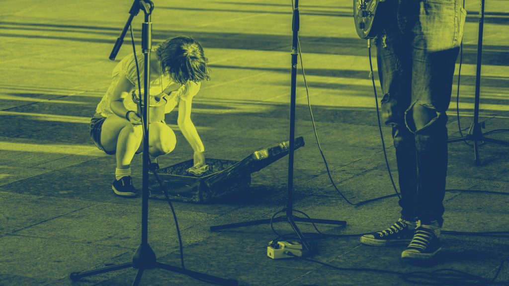 En person sitter på huk och lägger pengar i ett öppet gitarrfodral bredvid en gatumusikant med mikrofon.