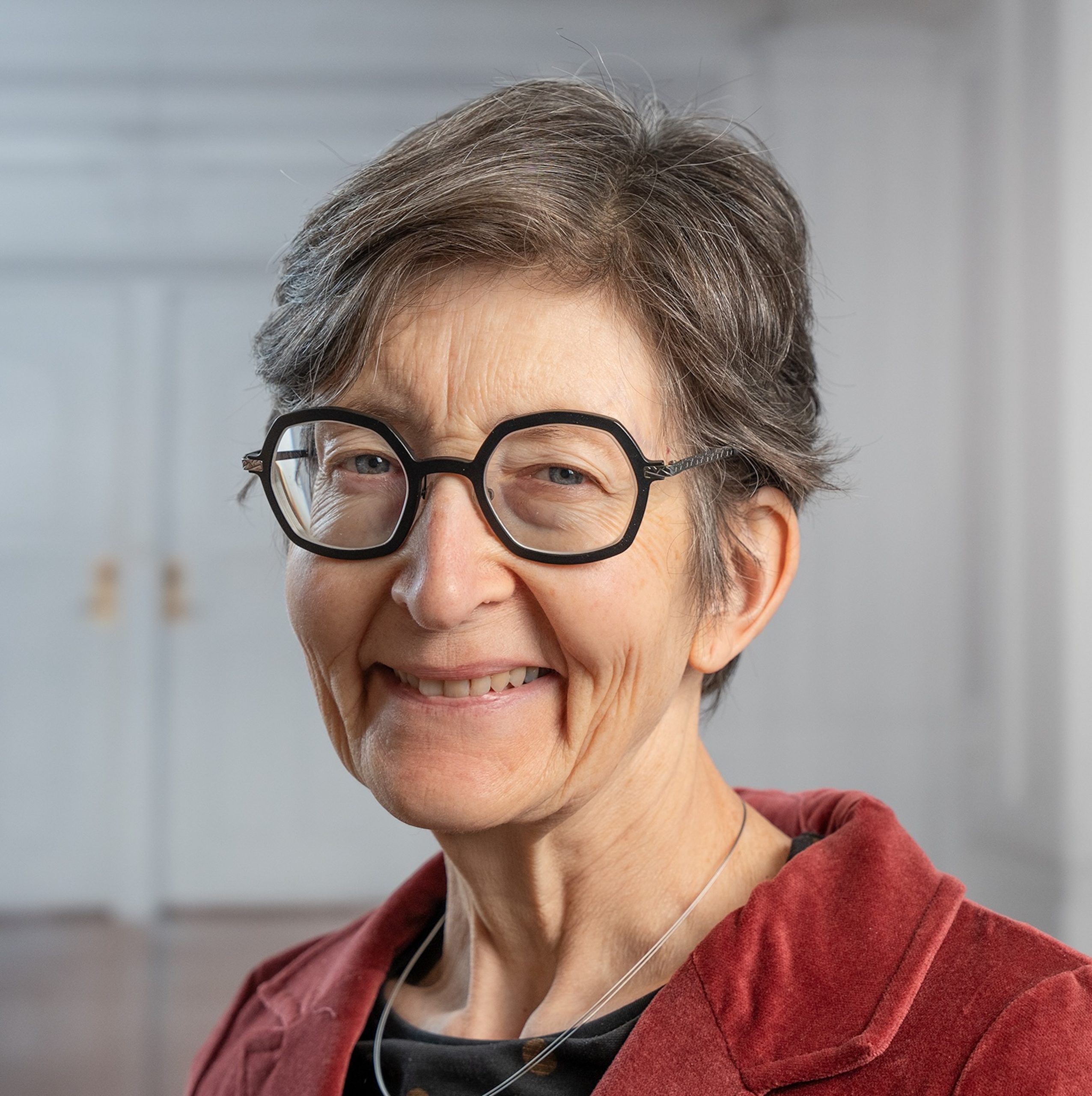 Leende äldre kvinna med kort grått hår, glasögon och röd jacka, stående inomhus.