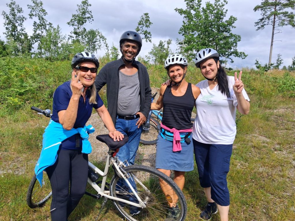 Fyra personer med hjälm poserar och ler vid sina cyklar utomhus en solig dag och gör fredstecken.