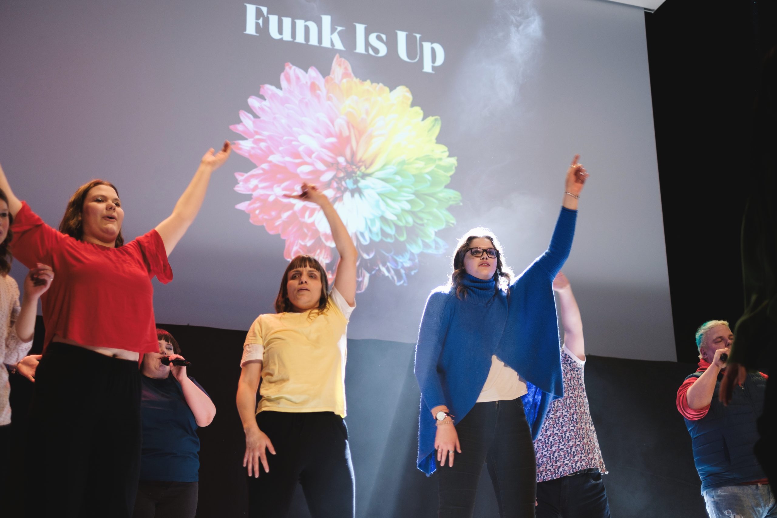 Grupp av människor på scen med armarna i luften; "Funk Is Up" och en färgglad blomma projiceras bakom dem.