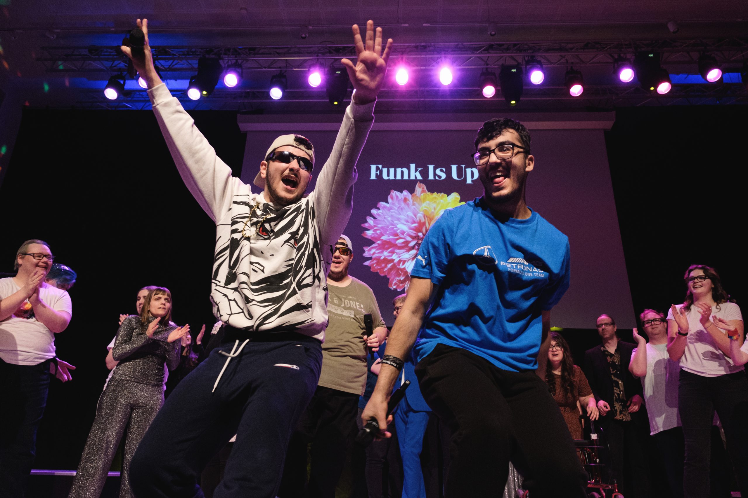 Två män dansar energiskt på scenen med en grupp, färgglada lampor och "Funk Is Up" projicerad bakom dem.