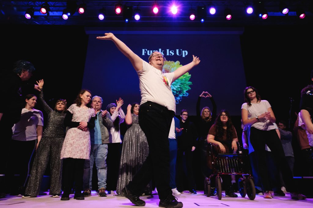 En glad grupp på scenen, med en person längst fram som intar en dramatisk, festlig pose under färgglada lampor.