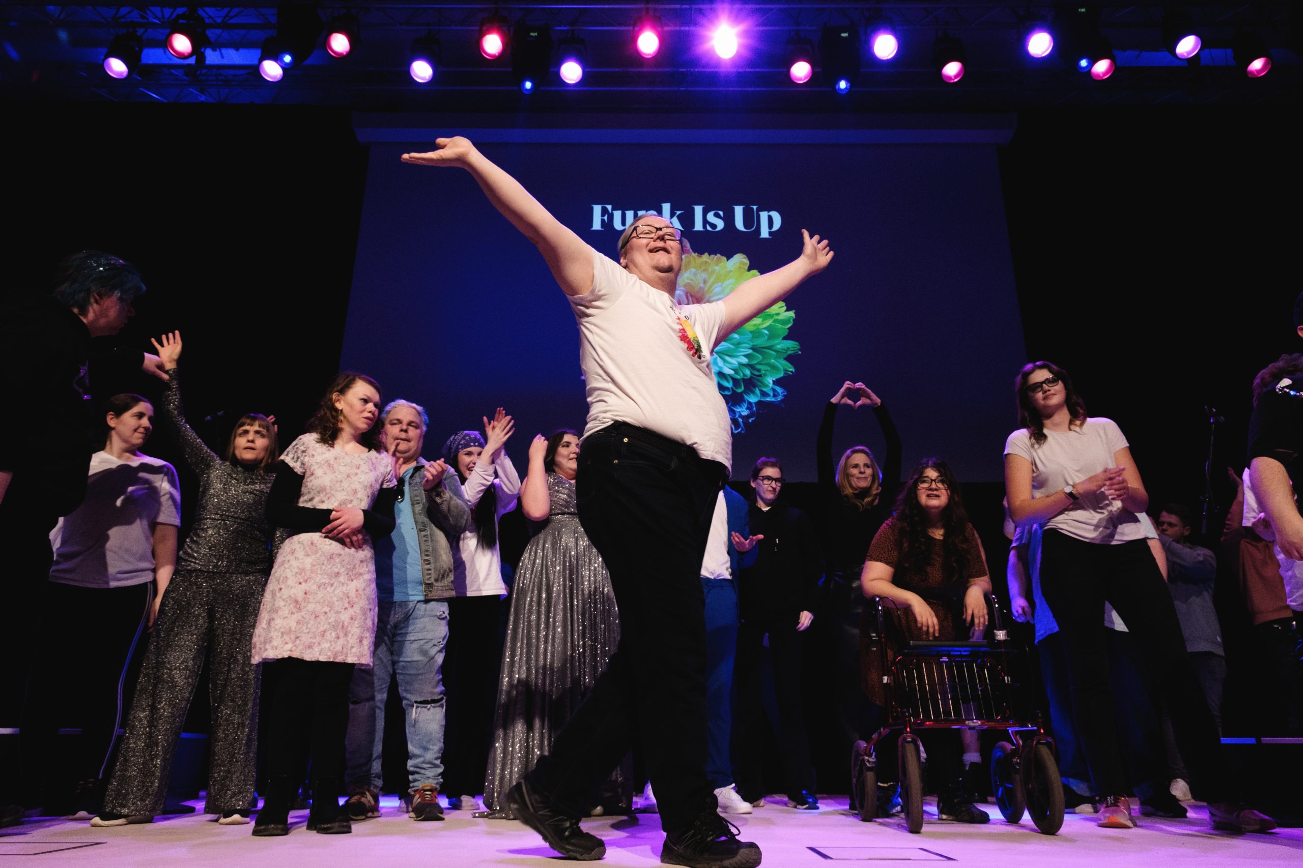 En glad grupp på scenen, med en person längst fram som intar en dramatisk, festlig pose under färgglada lampor.