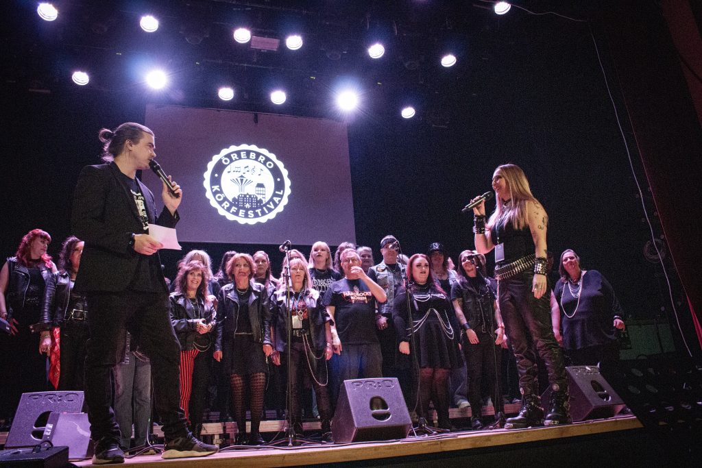 En grupp människor på scenen, några med mikrofoner, i starkt ljus med en festivallogotyp i bakgrunden.