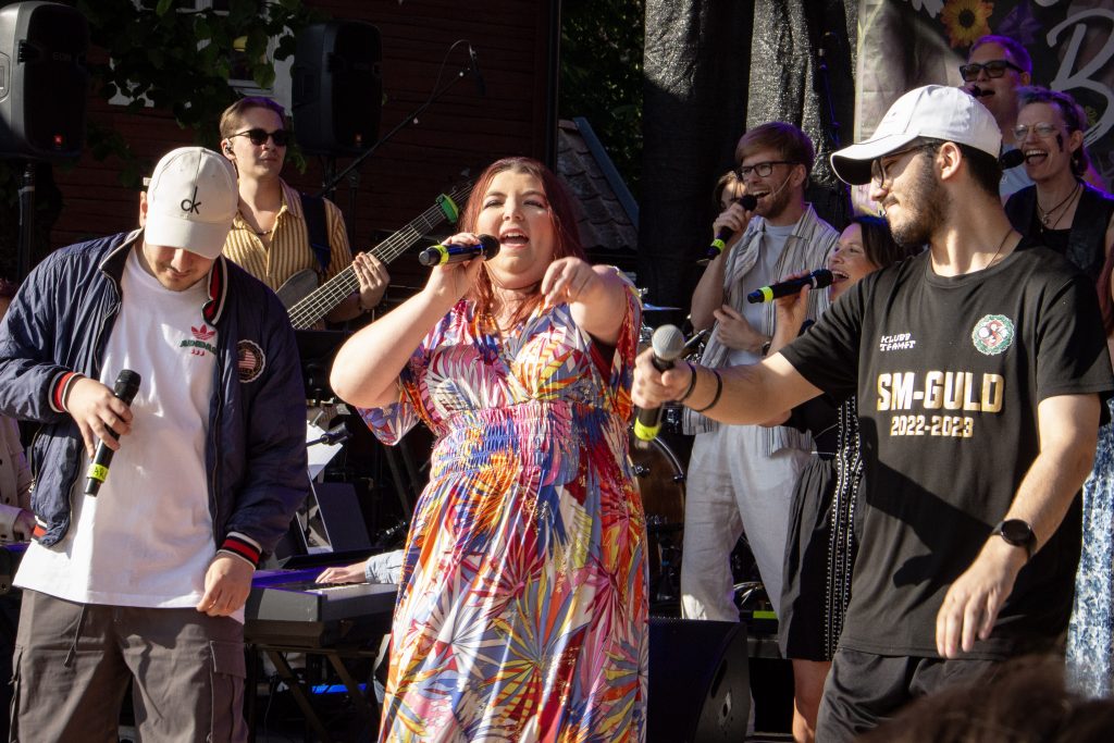 En grupp sångare uppträder energiskt på en utomhusscen med mikrofoner och musikinstrument.