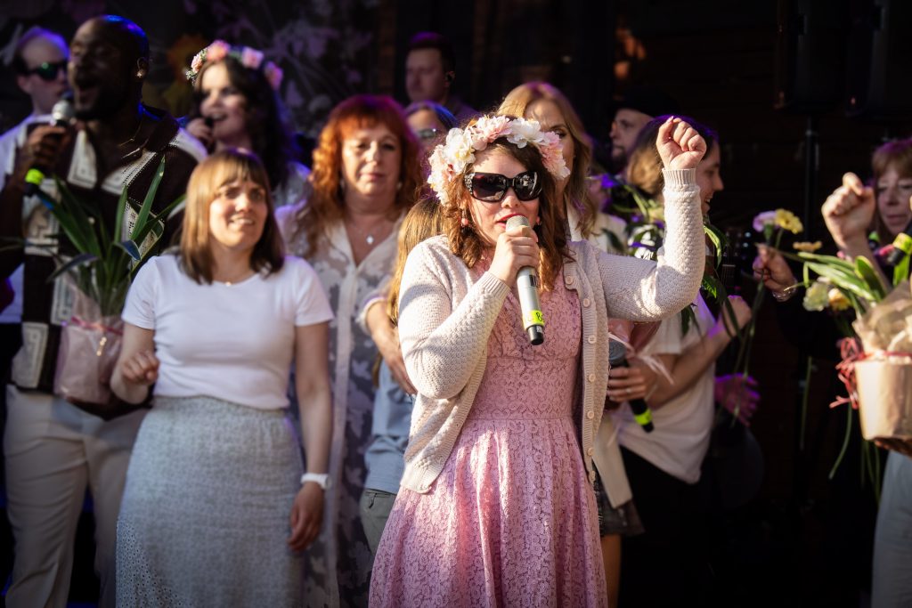 Kvinna i rosa klänning och blomsterkrona sjunger i mikrofon på scen med en grupp människor.