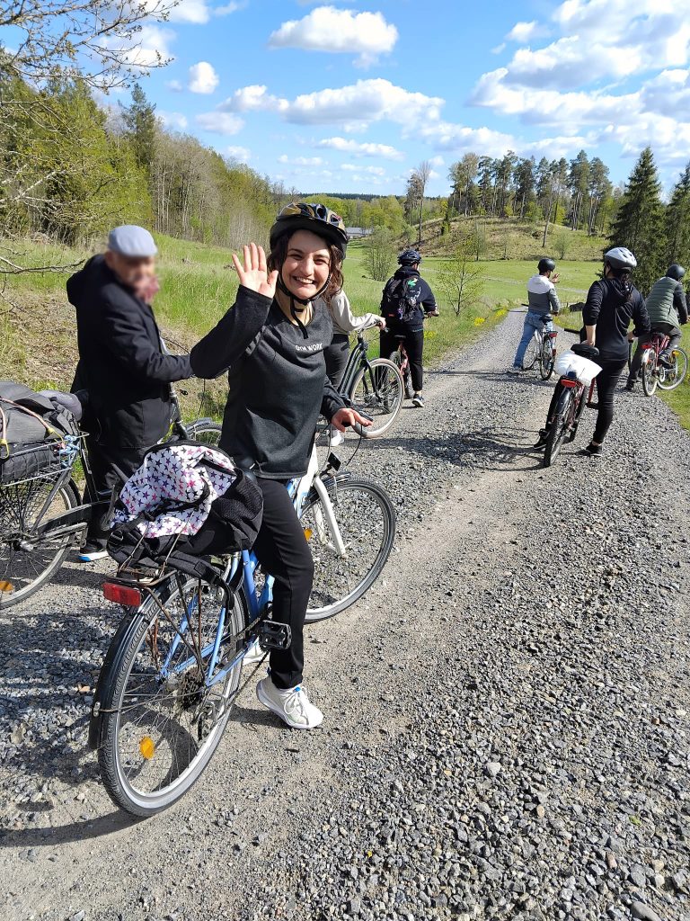 Leende kvinna på cykel vinkar medan andra med cyklar pausar på en naturskön grusväg under en blå himmel.