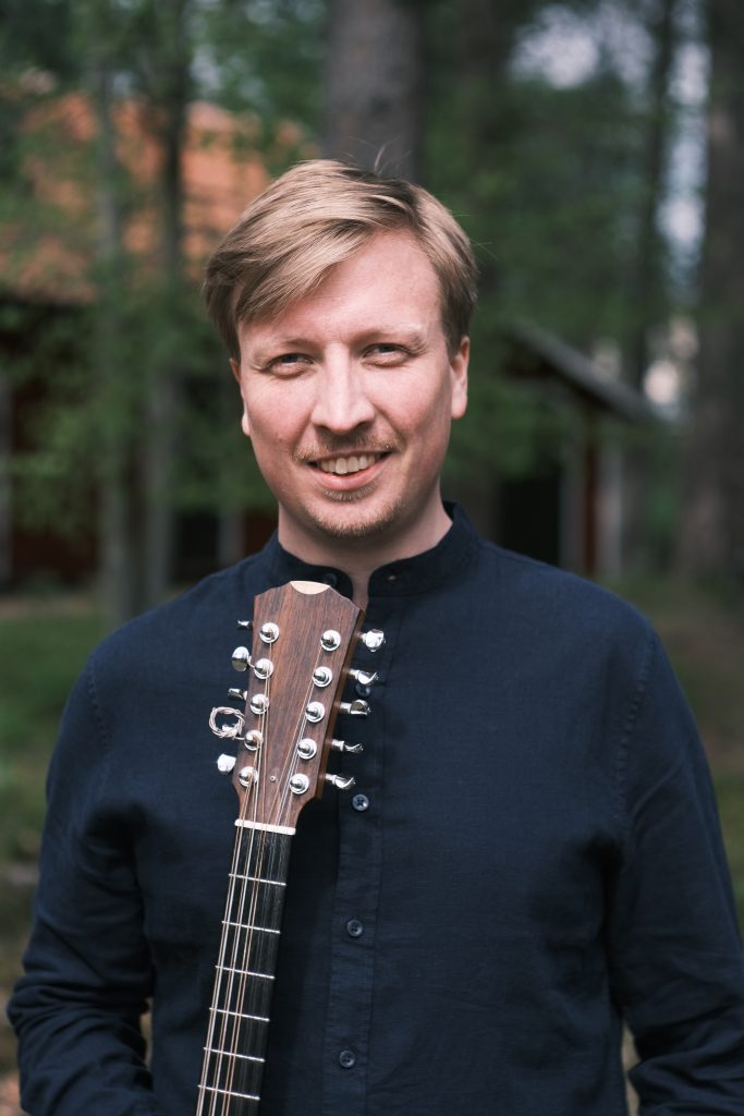 Leende man i mörk skjorta håller i en gitarr utomhus, med träd och byggnader suddiga i bakgrunden.
