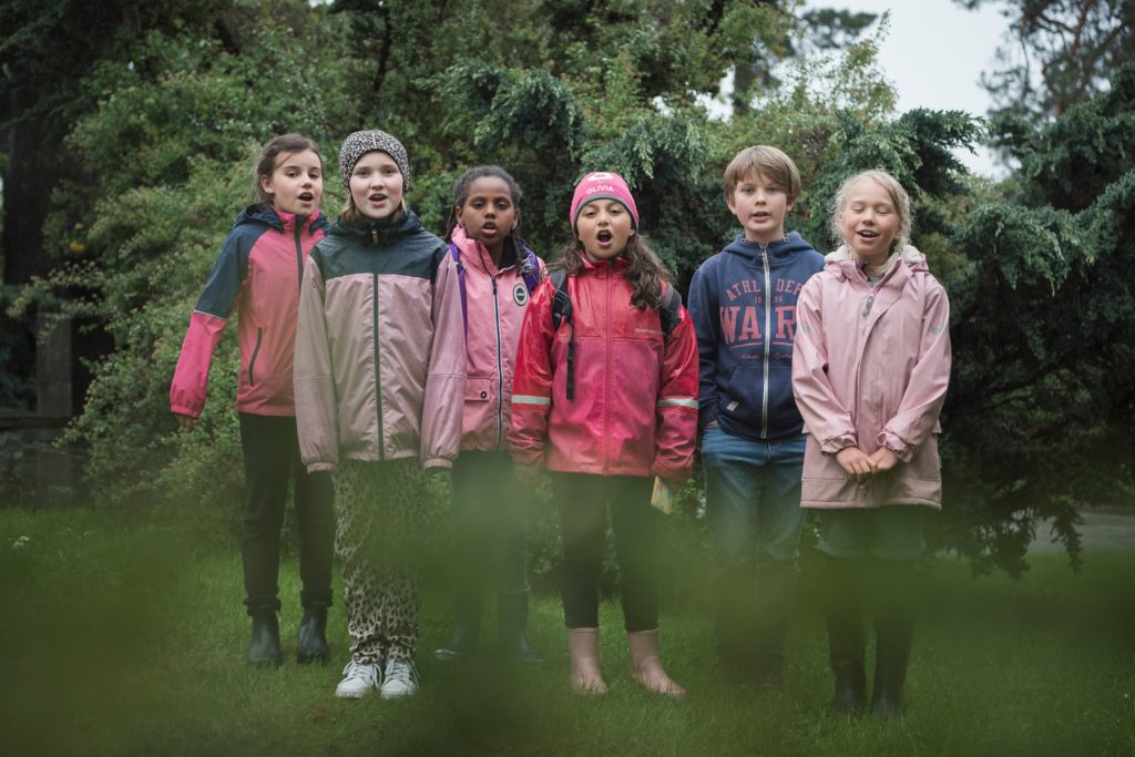 Sex barn i kavaj står på rad utomhus och sjunger tillsammans framför gröna träd.