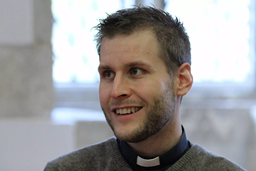 En leende man med kort hår och kontorskrage, inomhus med ett suddigt fönster i bakgrunden.