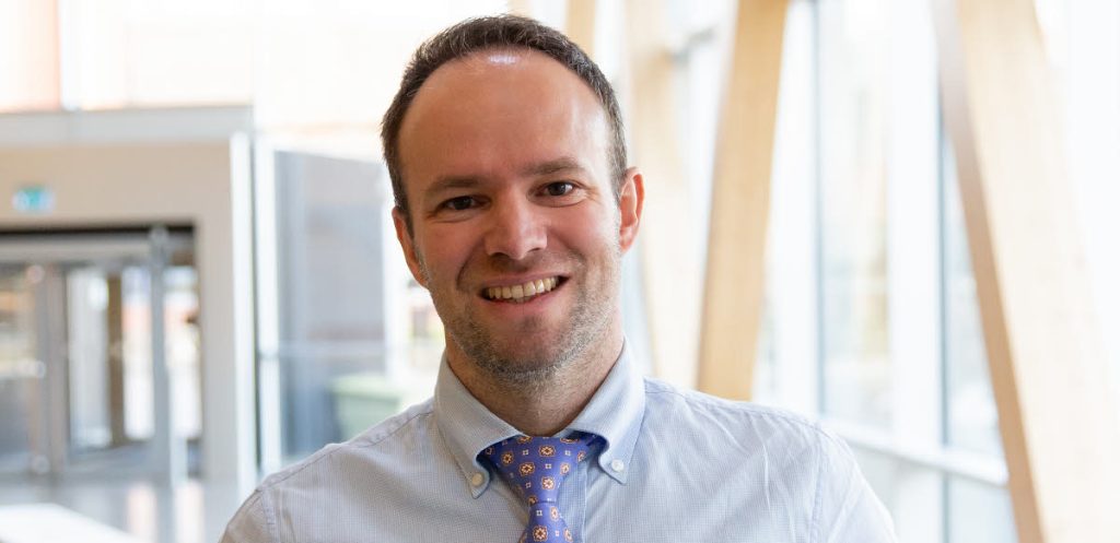 Leende man i blå skjorta och mönstrad slips står i en ljus, modern hall med stora fönster.