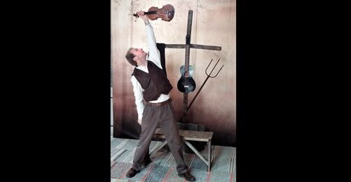 Man i vintagekläder står på tå och sträcker sig upp med en violin i handen; bakom honom finns bland annat en högaffel.