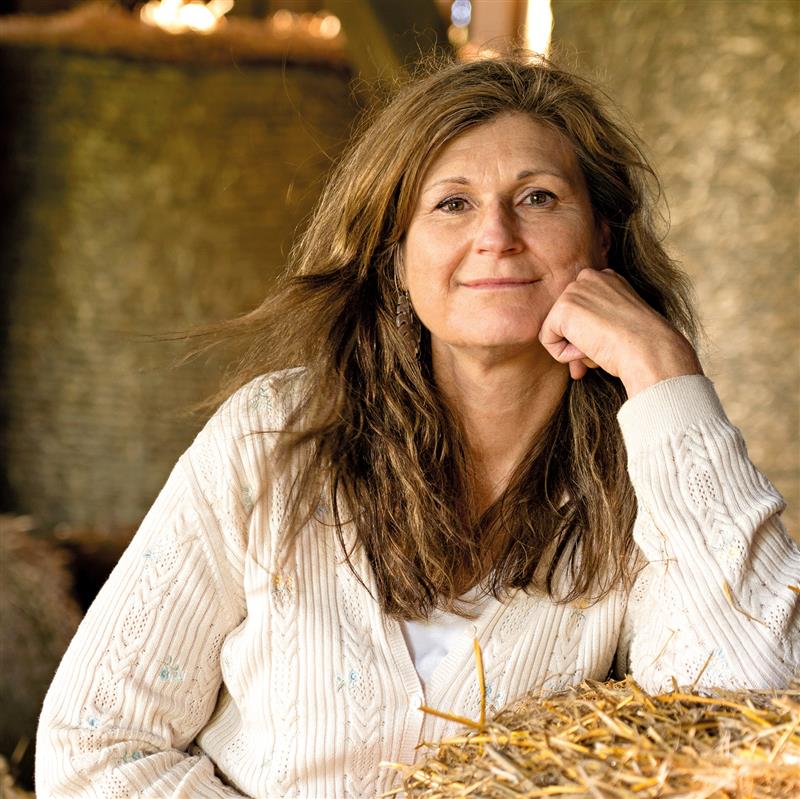 Kvinna med långt brunt hår i vit tröja, sitter vid hö och ler mot kameran.