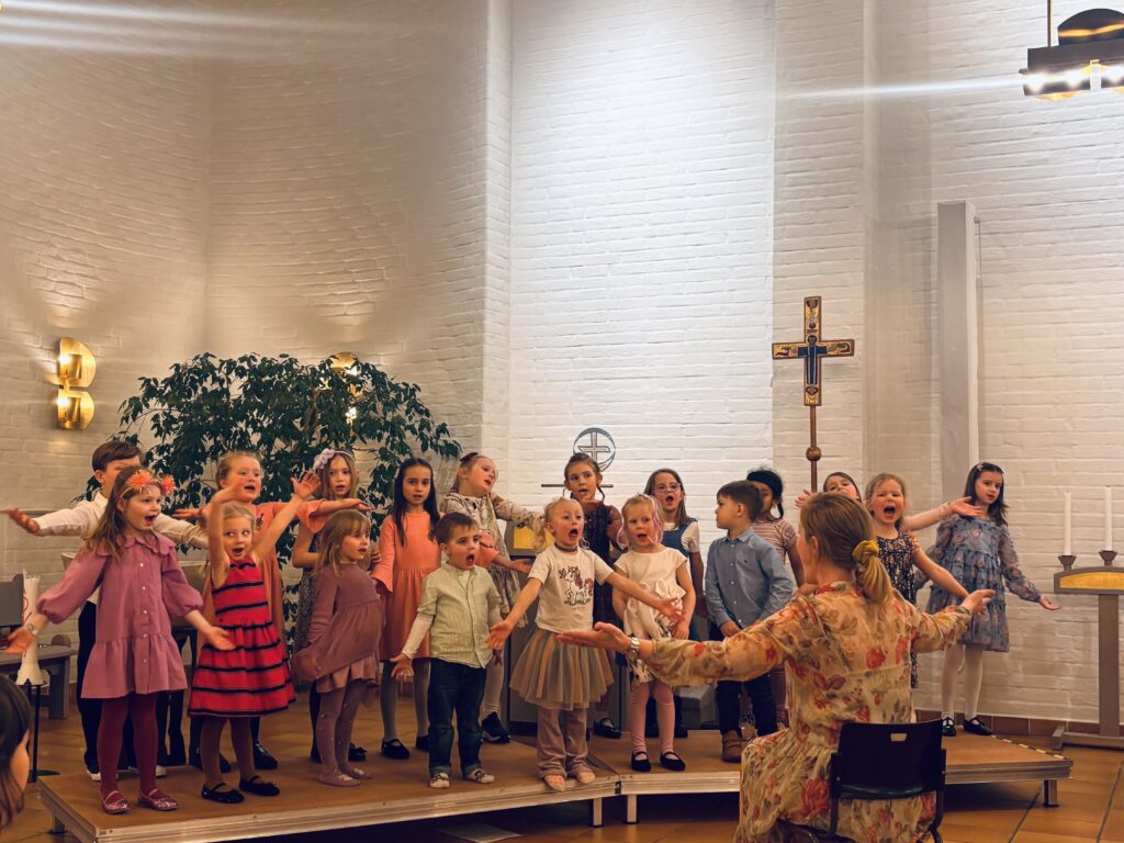 En grupp barn sjunger på scenen med en dirigent framför sig, i ett ljust rum med vita tegelväggar.