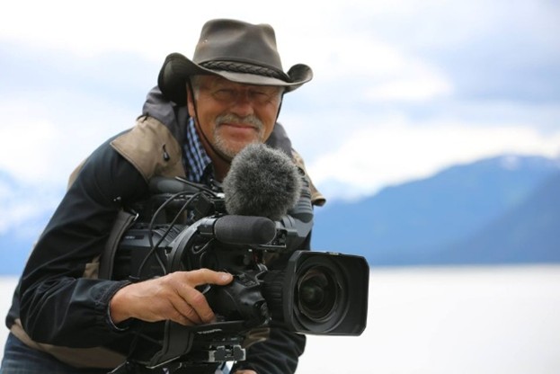 Man i hatt ler medan han använder en professionell videokamera utomhus med berg i bakgrunden.